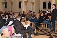 Eine Gruppe von Leuten in Kirchenbänken, die einem sprechenden Mann zuhört.